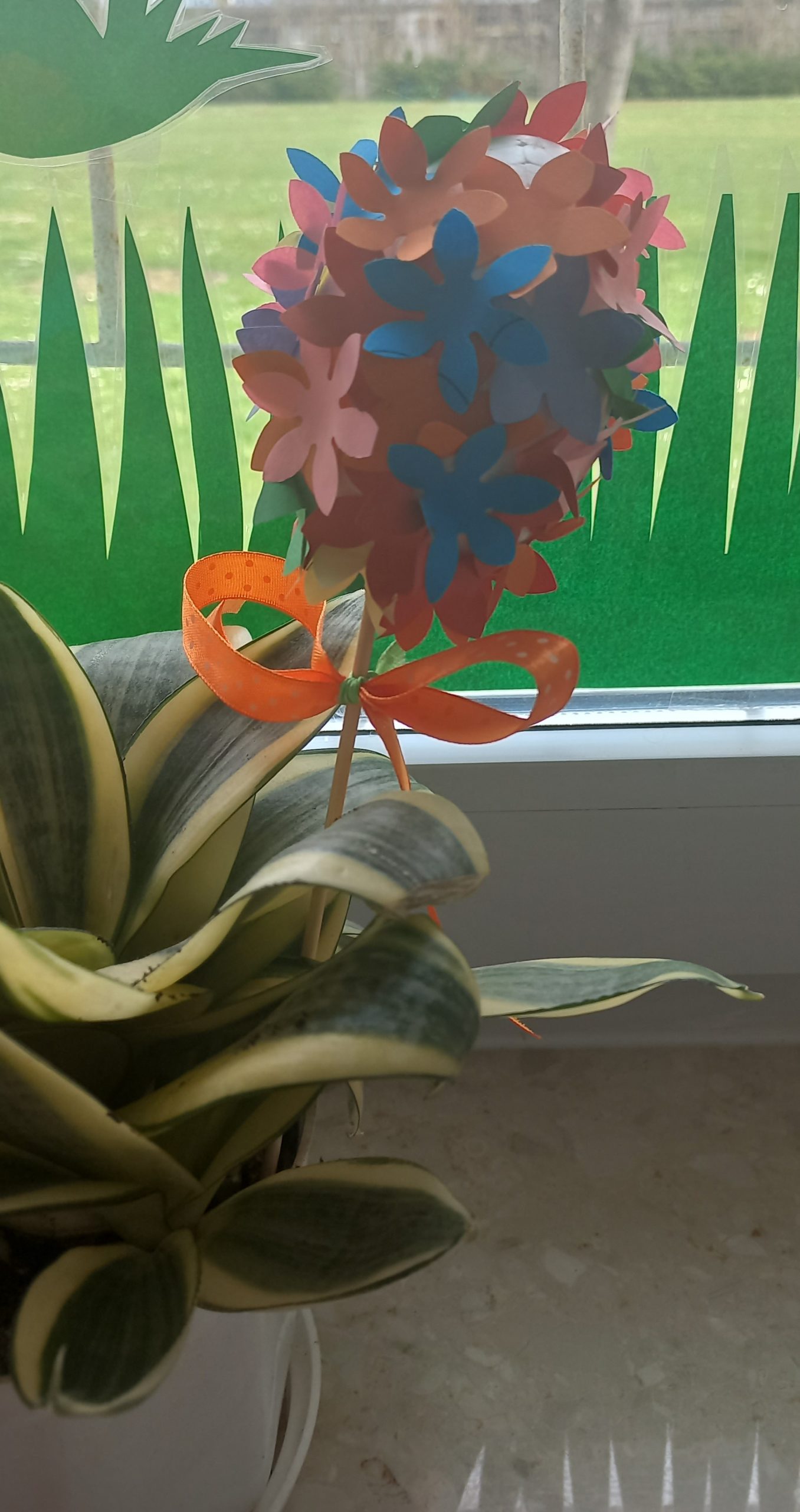 Doniczka z kwiatem w kształcie zielonych liści. W doniczce na długiej wykałaczce pisanka z kolorowymi kwiatami z papieru.