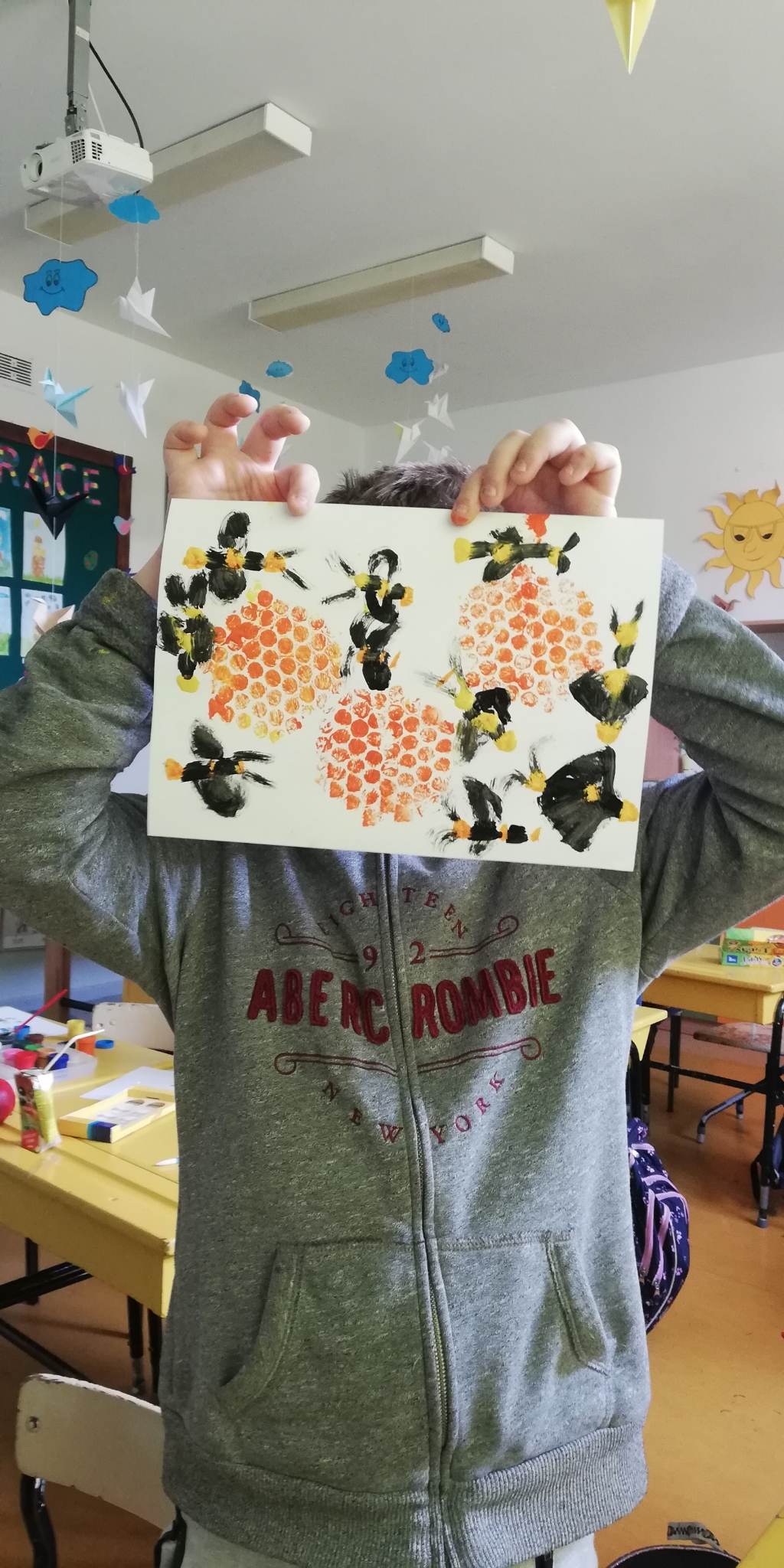 chłopiec zakrywa sobie twarz i jednocześnie pokazuje swój obrazek w pszczółki który namalował