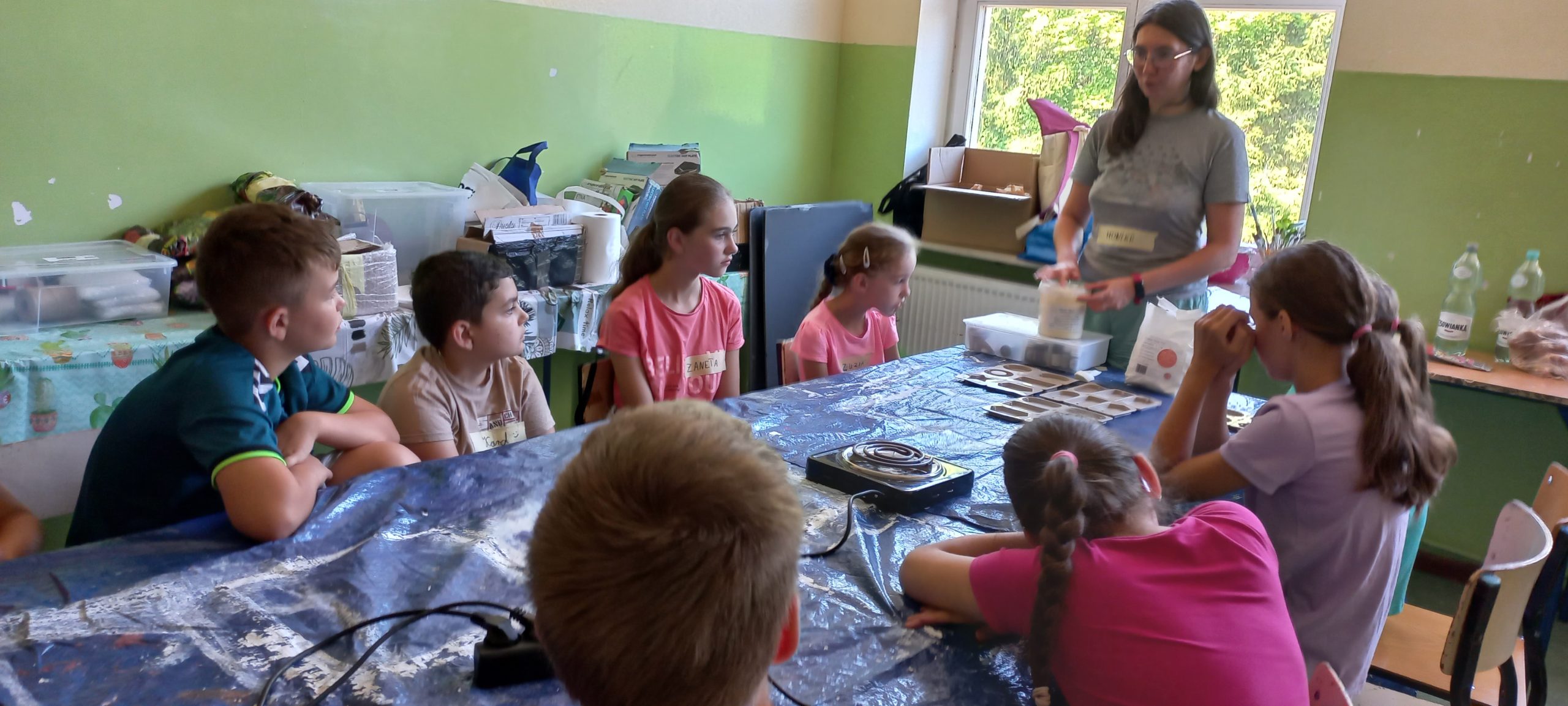 dzieci siedzą przy stole i słuchają prowadzącej, która tłumaczy proces tworzenia mydełek