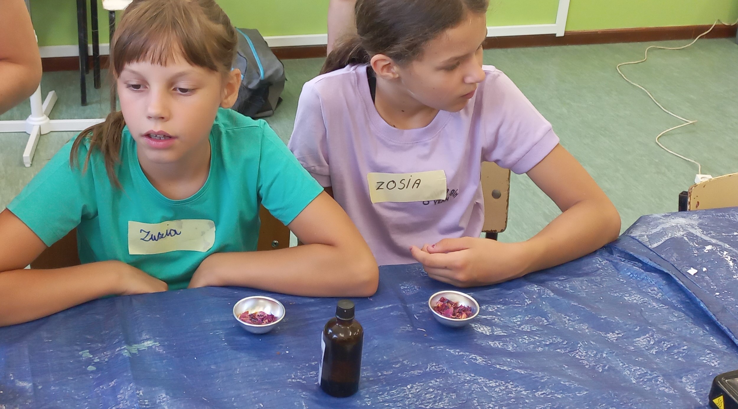 dwie dziewczynki siedzą przy stole na warsztatach z robienia mydełek, przed nimi na stole stoi buteleczka z jakimś płynem oraz suszone kwiatki