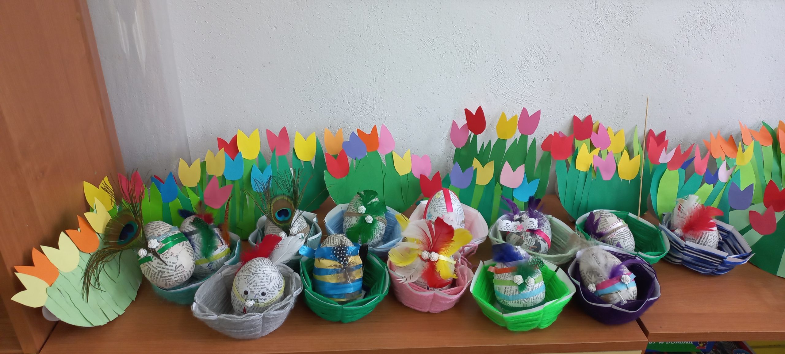 na tle kolorowych tulipanów z papieru widać małe koszyczki z włóczki z pisankami w środku
