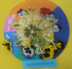 W środku kolorowej ramki wykonanej z talerzyka papierowego widoczne żywe kwiaty. Na dole ramki napis: Kochanej Mamusi