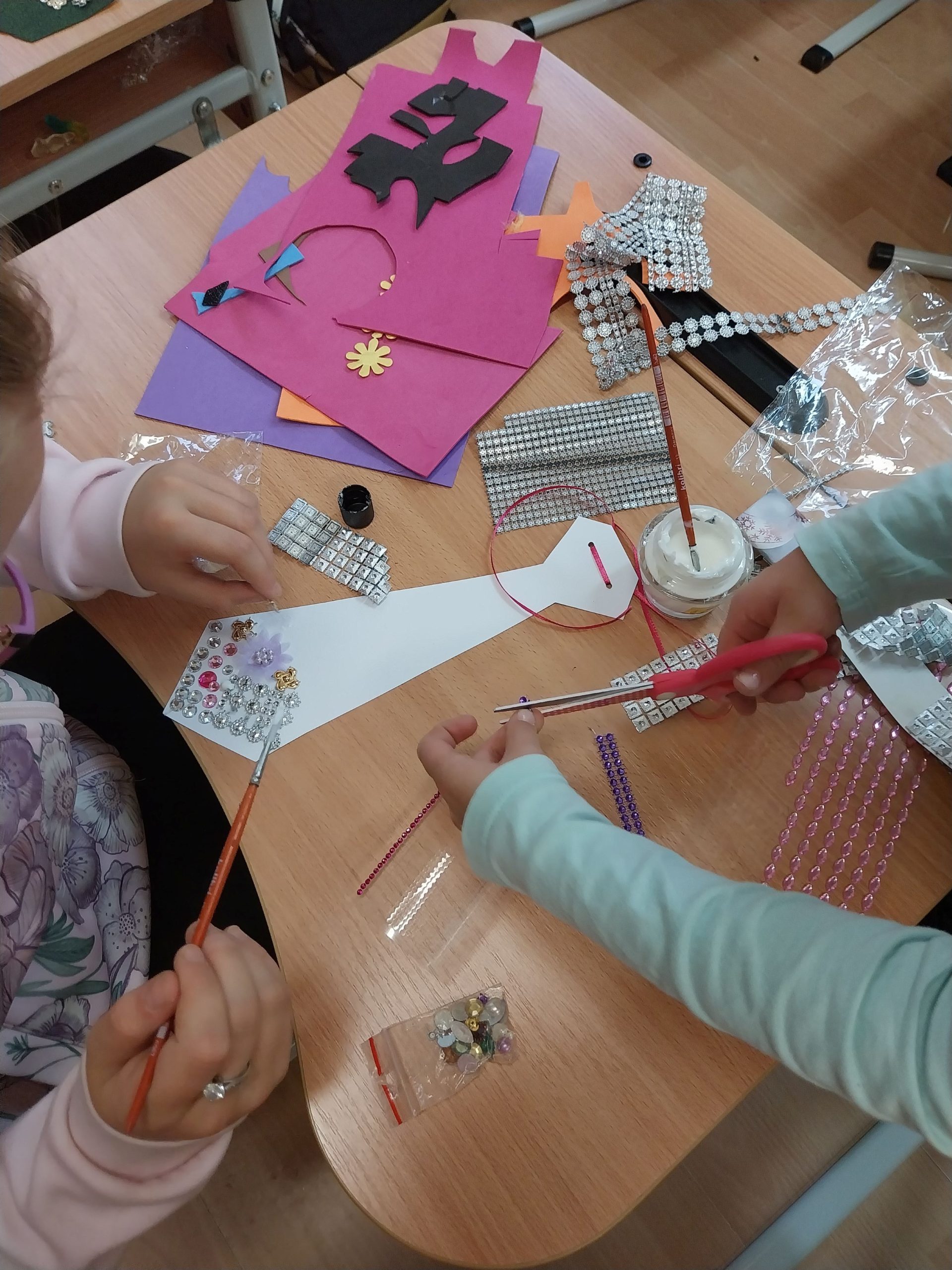 dwójka dzieci siedzi przy stole i przykleja kolorowe ornamenty dekoracyjne na kartonowe krawaty