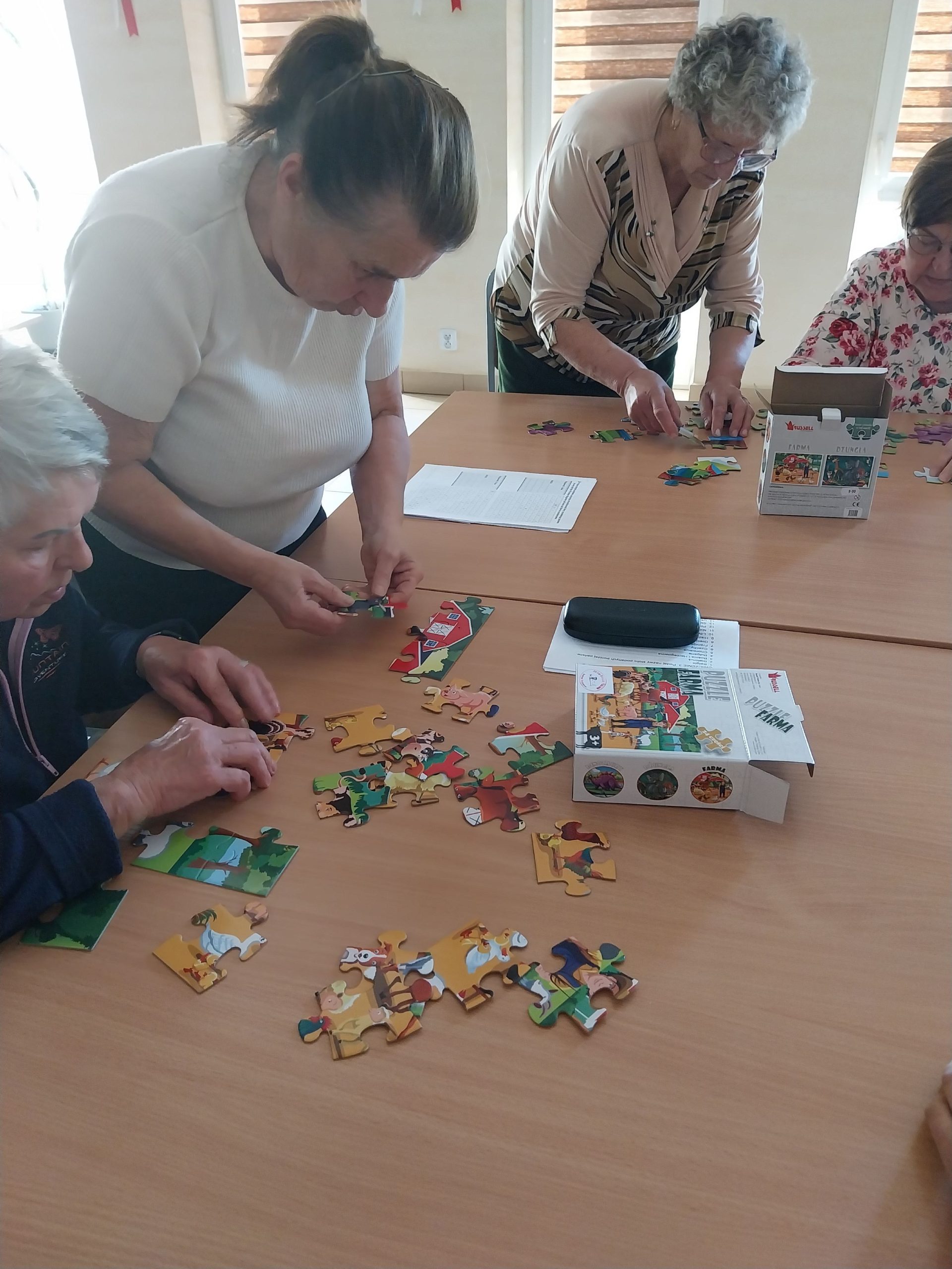 przy stoliku trzy Panie układają puzzle analogowe. Jedna z nich stoi, dwie siedzą na krzesłach