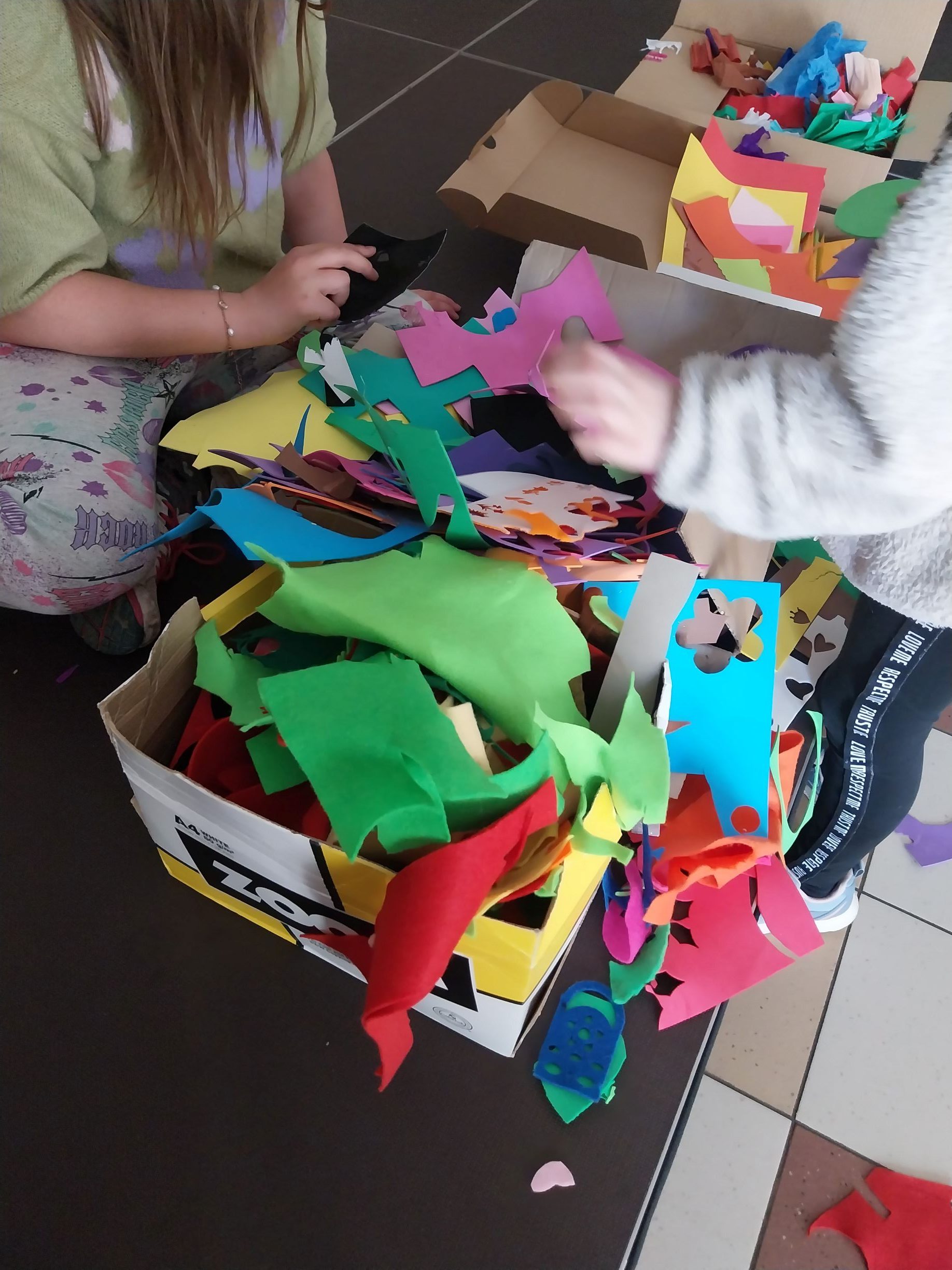 dwie dziewczynki przeszukują pudła z materiałami plastycznymi celem dobrania koloru swojej pracy