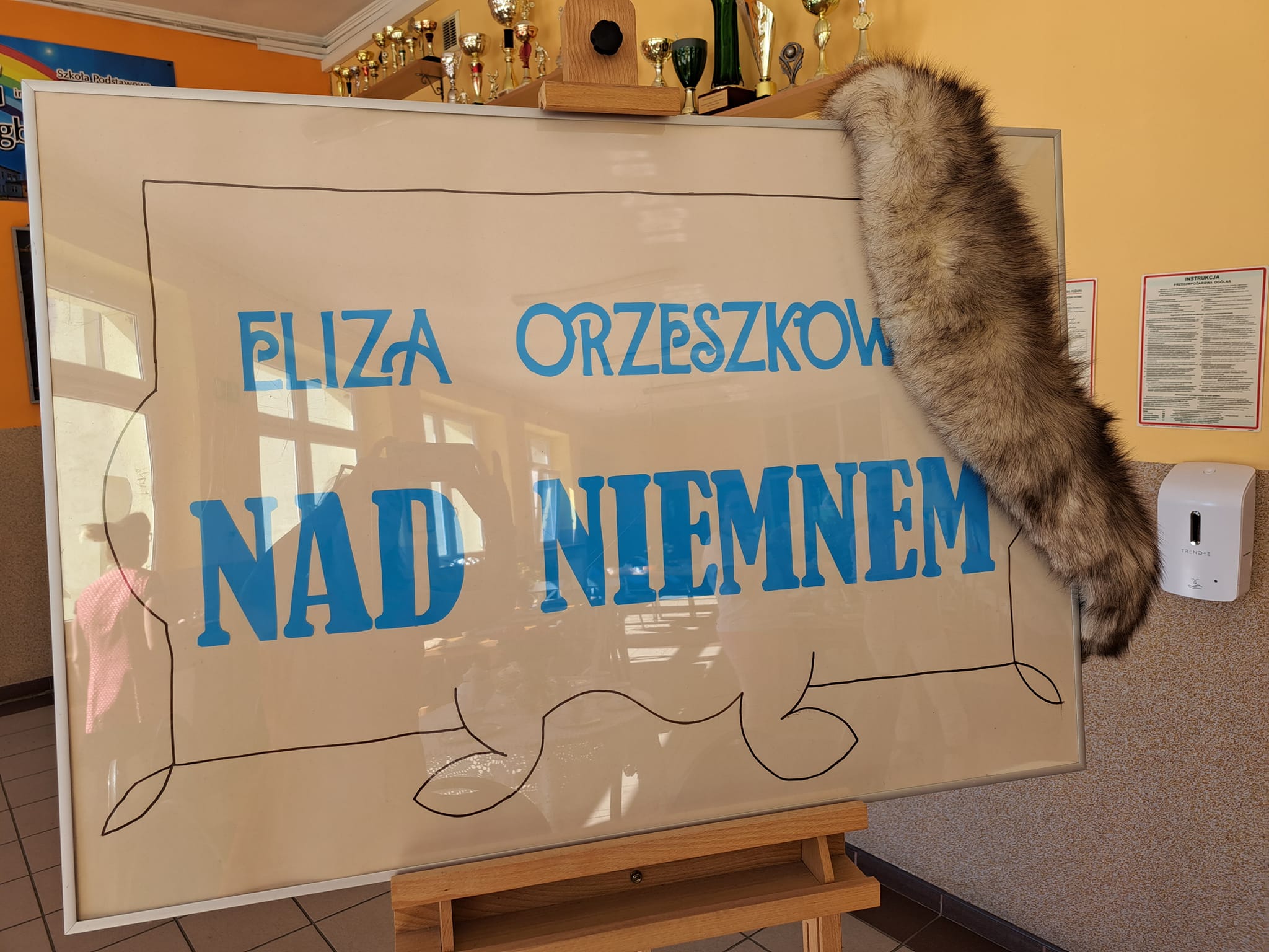 Kolorowe zdjęcie. Na sztaludze rama z plakatem z napisem Eliza Orzeszkowa Nad Niemnem. Na ramie futrzasty szal.